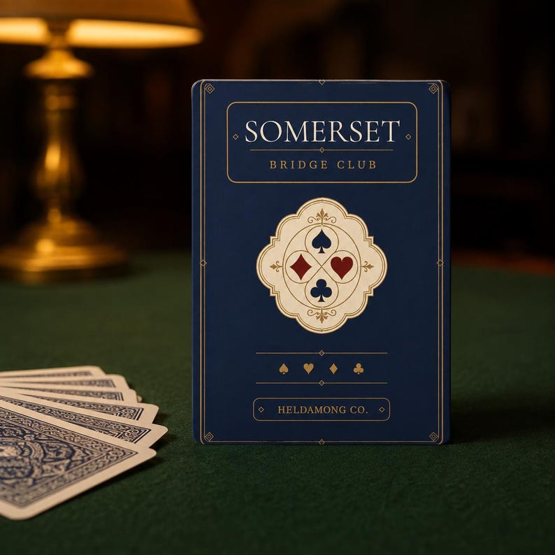 Puede incluir: Una baraja de cartas rectangular en una caja azul oscuro con letras doradas. La caja dice SOMERSET BRIDGE CLUB y HELDAMONG CO. Un emblema central de color crema muestra los cuatro palos de la baraja en rojo y azul, con iconos m&aacute;s peque&ntilde;os.