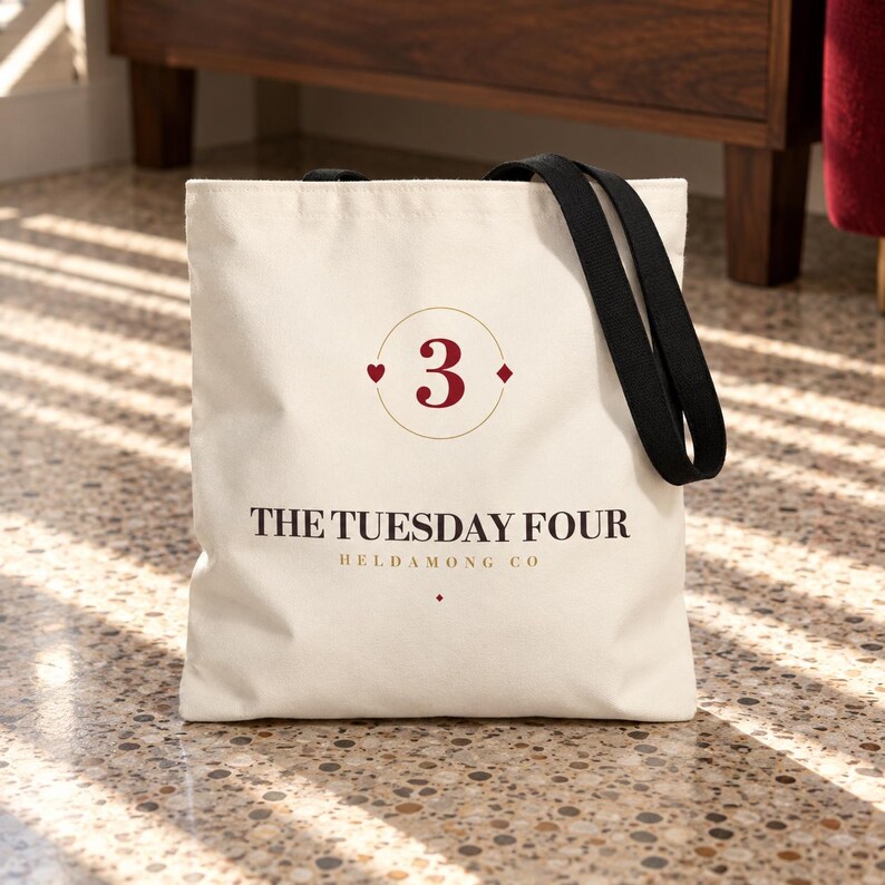 Puede incluir: Un bolso de tela color crema con dos asas de tela negra. La parte frontal presenta un n&uacute;mero 3 rojo oscuro dentro de un c&iacute;rculo dorado fino, flanqueado por un coraz&oacute;n y un diamante. Debajo, el texto dice THE TUESDAY FOUR y HELDAMONG CO.