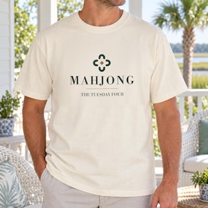 May include: A cream-coloured short-sleeve t-shirt featuring a dark green graphic on the chest. The design includes a stylised four-petal symbol above the text "MAHJONG" and the smaller text "THE TUESDAY FOUR" centred below the main word.