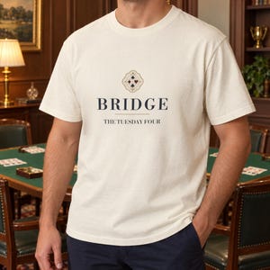 May include: An off-white short-sleeve t-shirt featuring a graphic print on the chest. The design includes a decorative emblem with four card suit symbols above the text BRIDGE and the smaller text THE TUESDAY FOUR in a dark serif font.