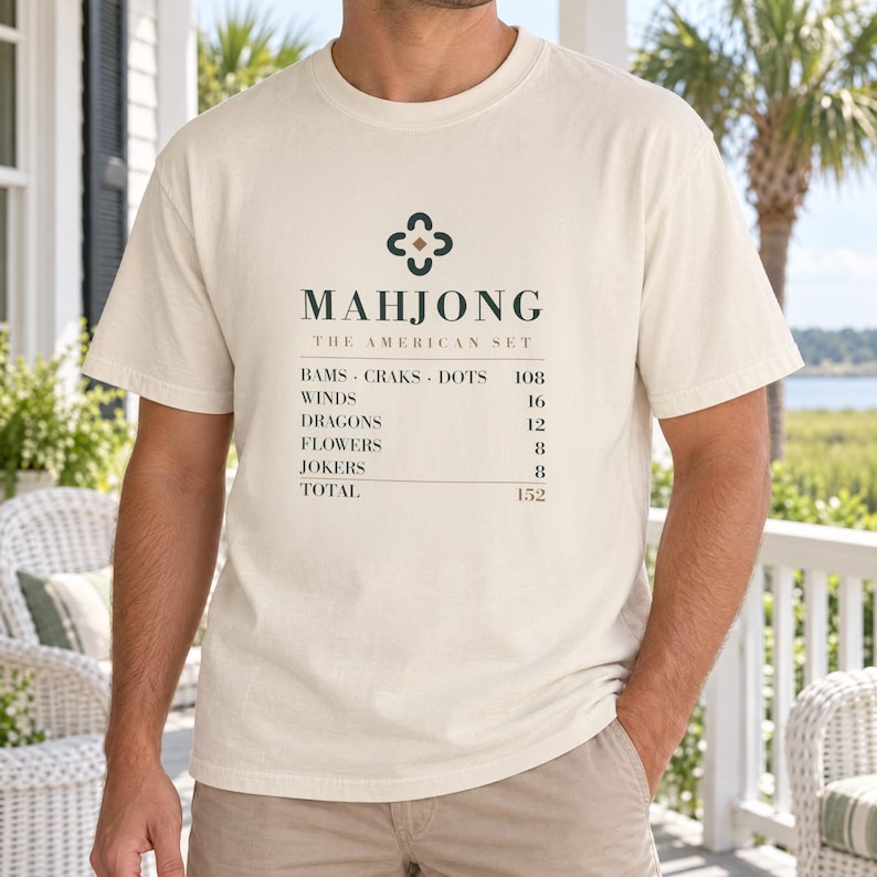 May include: A cream-colored short-sleeve t-shirt featuring a dark green graphic of a Mahjong symbol and the text: MAHJONG THE AMERICAN SET, followed by a list of tiles including BAMS, CRAKS, DOTS, WINDS, DRAGONS, FLOWERS, JOKERS, and a total of 152.