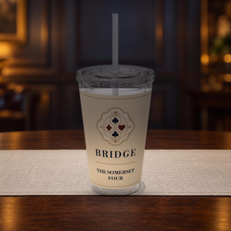 May include: A clear plastic tumbler with a lid and straw, featuring a cream-colored label. The label displays a decorative emblem with four card suit symbols, the word BRIDGE, and the text THE SOMERSET FOUR in black, centered on the cup.