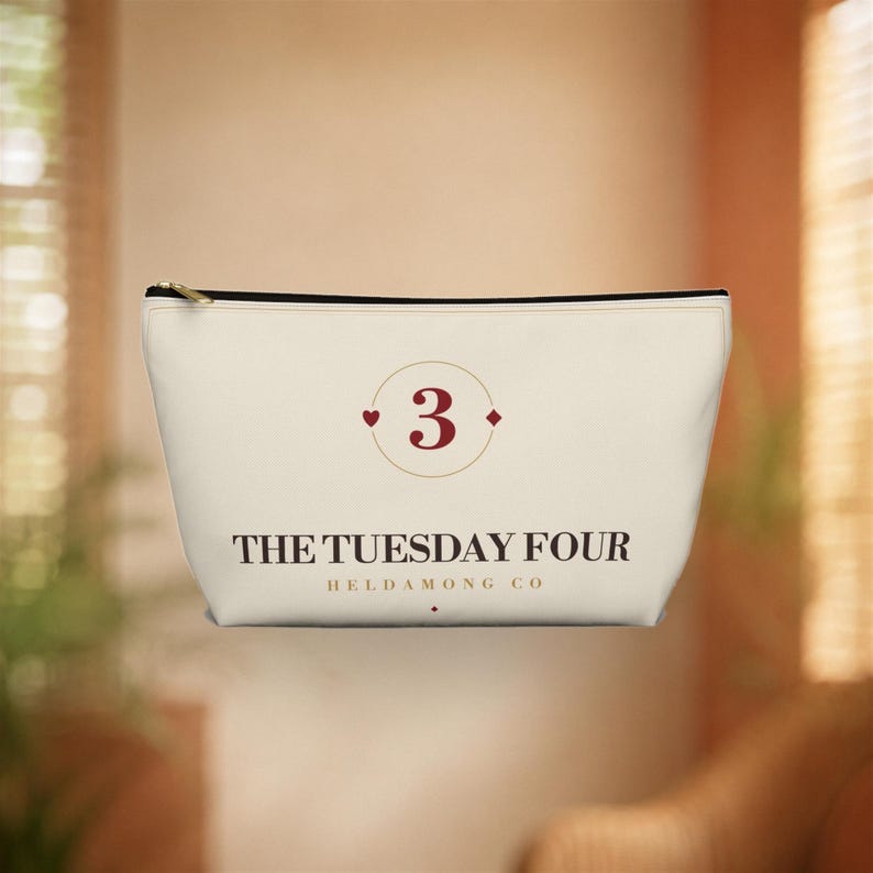 May include: A cream-colored zippered pouch featuring a centered dark red number 3 enclosed in a thin circular frame. Below the number, the text reads THE TUESDAY FOUR in a bold serif font, with HELDAMONG CO printed in smaller letters underneath.