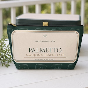 以下が含まれることがあります： HELDAMONG CO、PALMETTO MAHJONG ESSENTIALS、MADE FOR YOUR PEOPLEという文字が記されたクリーム色の長方形ラベルが付いたダークグリーンのファスナー付きポーチ。ポーチには、明るい緑色の麻雀牌の輪郭が繰り返されるパターンが施されています。