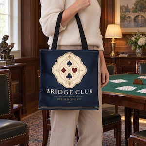 May include: A navy blue tote bag with black shoulder straps featuring a cream-colored ornate emblem containing the four card suit symbols. The text BRIDGE CLUB is printed in white capital letters above the text HELDAMONG CO. in smaller font.