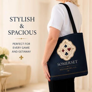 May include: A dark blue tote bag with black shoulder straps featuring a cream-coloured emblem with four playing card suit symbols. The text reads STYLISH & SPACIOUS, PERFECT FOR EVERY GAME AND GETAWAY, SOMERSET, and HELDAMONG CO. with a fleur-de-lis.