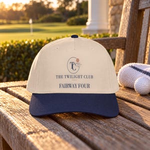 May include: A cream-colored baseball cap with a dark navy blue brim. The front features a logo with the letters TC inside an oval, a small circle, and the text THE TWILIGHT CLUB and FAIRWAY FOUR printed in dark navy blue lettering.