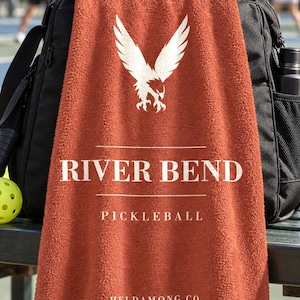 May include: A rust-colored towel featuring a white graphic of an eagle with wings spread above the text RIVER BEND, PICKLEBALL, and HELDAMONG CO. The towel is draped over a black sports bag, with a yellow pickleball visible on the left side.