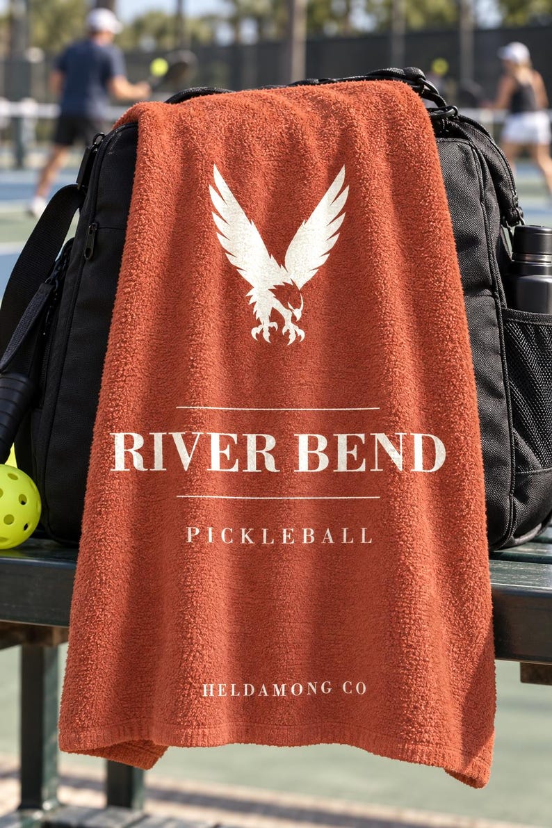 May include: A rust-colored towel featuring a white graphic of an eagle with wings spread above the text RIVER BEND, PICKLEBALL, and HELDAMONG CO. The towel is draped over a black sports bag, with a yellow pickleball visible on the left side.