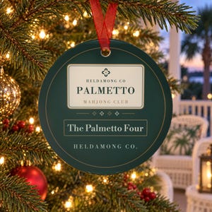 May include: A round dark green ornament with a red ribbon hanger. The center features a cream-colored label with the text HELDAMONG CO PALMETTO MAHJONG CLUB, followed by the text The Palmetto Four and the brand name HELDAMONG CO. at the bottom.
