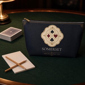 Peut inclure: Une pochette zipp&eacute;e bleu marine fonc&eacute; orn&eacute;e d'un embl&egrave;me cr&egrave;me repr&eacute;sentant quatre symboles de cartes &agrave; jouer : pique, carreau, c&oelig;ur et tr&egrave;fle. Le texte SOMERSET et HELDAMONG CO. est imprim&eacute; en blanc sous l'embl&egrave;me, avec des bordures dor&eacute;es.