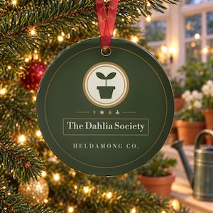 May include: A round, dark green ceramic ornament featuring a central cream-colored circle with a potted plant icon. Below the icon, the text reads The Dahlia Society and HELDAMONG CO. in white lettering. A red ribbon is attached to the top for hanging.