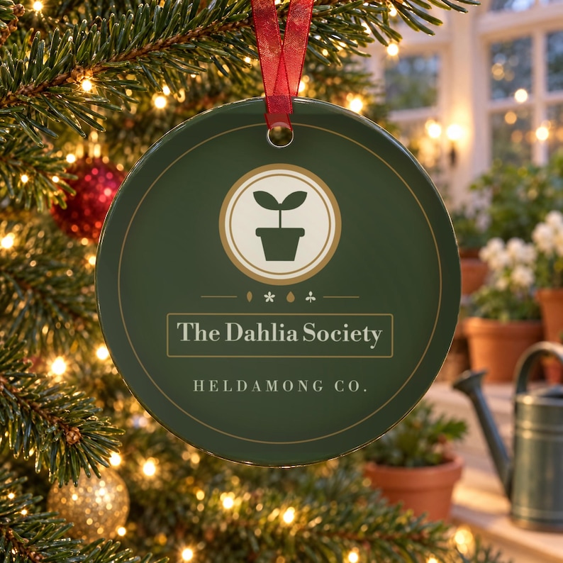 May include: A round, dark green ceramic ornament featuring a central cream-colored circle with a potted plant icon. Below the icon, the text reads The Dahlia Society and HELDAMONG CO. in white lettering. A red ribbon is attached to the top for hanging.