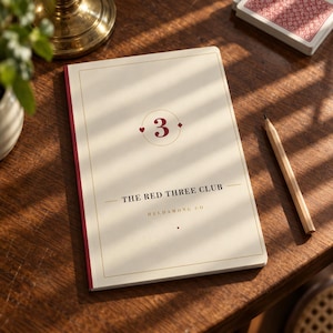 May include: A cream-colored notebook with a red spine featuring the number 3 inside a circle, the text THE RED THREE CLUB, and HELDAMONG CO. The cover has a thin gold border and a small red diamond and heart icon flanking the number 3.