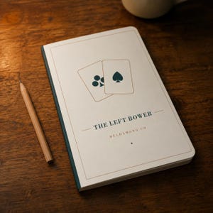 May include: A rectangular notebook with a white cover featuring a graphic of two playing cards, one with a club and one with a spade. The text reads THE LEFT BOWER and HELDAMONG CO. A wooden pencil lies beside the notebook on a dark wooden surface.