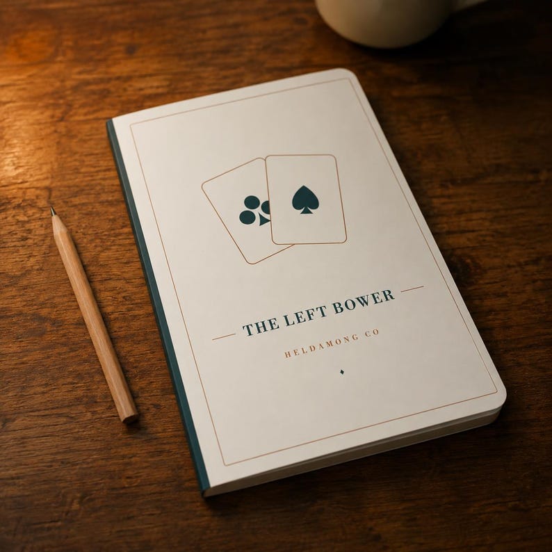 May include: A rectangular notebook with a white cover featuring a graphic of two playing cards, one with a club and one with a spade. The text reads THE LEFT BOWER and HELDAMONG CO. A wooden pencil lies beside the notebook on a dark wooden surface.