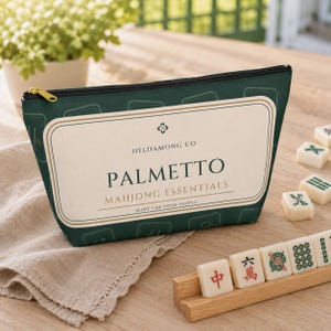 May include: A dark green zippered pouch featuring a cream-colored rectangular label with the text HELDAMONG CO, PALMETTO, MAHJONG ESSENTIALS, and MADE FOR YOUR PEOPLE. The pouch has a repeating pattern of mahjong tile outlines.