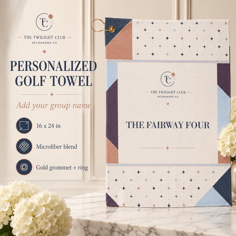 May include: A rectangular microfiber golf towel featuring a geometric pattern in navy blue, light blue, and muted peach. The center displays the text THE TWILIGHT CLUB HELDAMONG CO and THE FAIRWAY FOUR. It includes a gold grommet and metal ring.