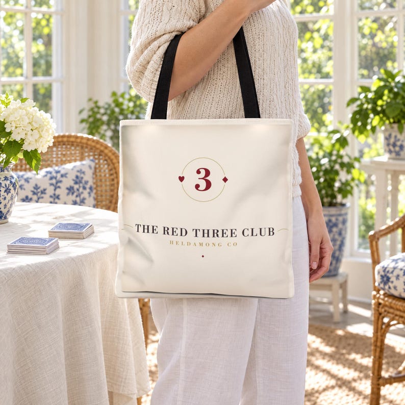 May include: A cream-coloured tote bag with black shoulder straps. The front features a dark red number 3 inside a circle, flanked by a small heart and diamond symbol. Below, the text reads THE RED THREE CLUB and HELDAMONG CO in black.