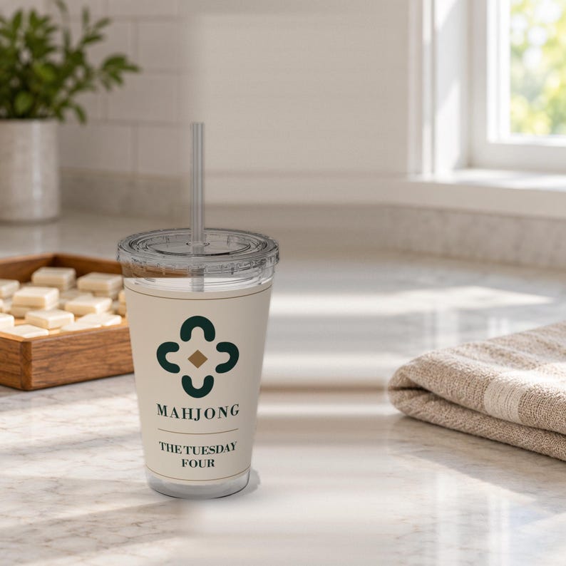 May include: A clear plastic tumbler with a lid and straw, featuring a cream-colored wrap with a dark green four-leaf clover-like graphic, a small gold diamond, and the black text "MAHJONG" followed by "THE TUESDAY FOUR" printed on the front.