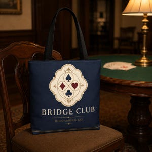 May include: A navy blue tote bag with black shoulder straps. The front features a cream-colored ornate emblem containing four card suit symbols: spade, heart, diamond, and club. Below the emblem, the text reads BRIDGE CLUB and HELDAMONG CO.