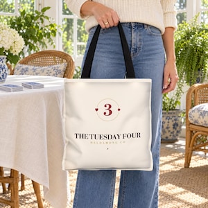 Puede incluir: Un bolso tote de color crema con asas negras que presenta el n&uacute;mero 3 en rojo oscuro dentro de un marco circular fino con peque&ntilde;os detalles de corazones y diamantes. Debajo, el texto THE TUESDAY FOUR HELDAMONG CO en letras marr&oacute;n oscuro.