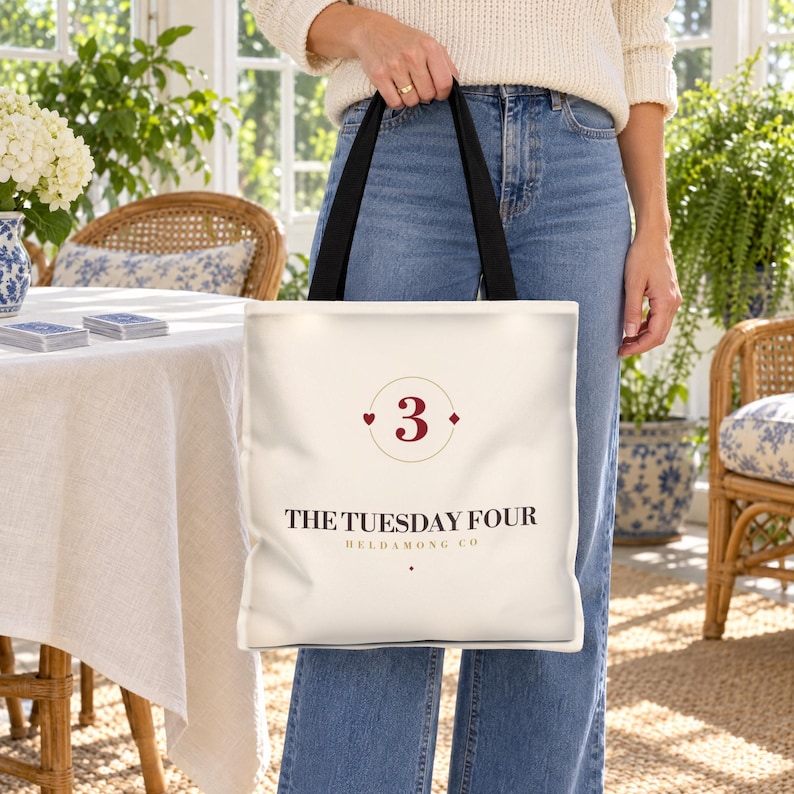 Puede incluir: Un bolso tote de color crema con asas negras que presenta el n&uacute;mero 3 en rojo oscuro dentro de un marco circular fino con peque&ntilde;os detalles de corazones y diamantes. Debajo, el texto THE TUESDAY FOUR HELDAMONG CO en letras marr&oacute;n oscuro.