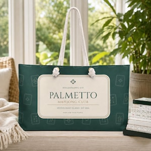 Pode incluir: Uma bolsa tote verde-escura com al&ccedil;as de corda branca e um padr&atilde;o repetitivo de pe&ccedil;as de mahjong. Uma etiqueta retangular cor de creme ao centro apresenta o texto: HELDAMONG CO, PALMETTO MAHJONG CLUB, HILTON HEAD ISLAND - EST 2026, MADE FOR YOUR PEOPLE.