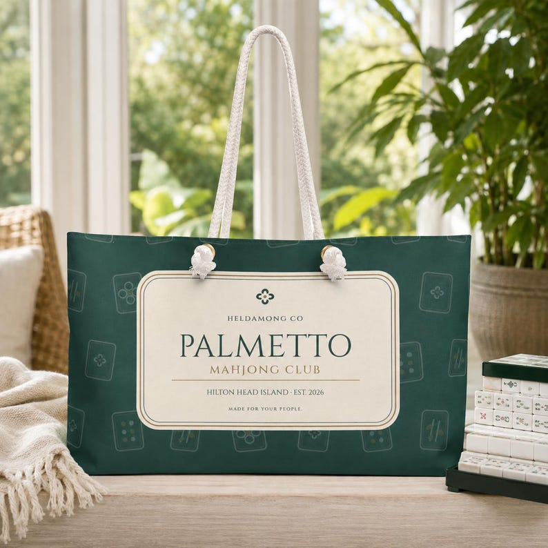 Pode incluir: Uma bolsa tote verde-escura com al&ccedil;as de corda branca e um padr&atilde;o repetitivo de pe&ccedil;as de mahjong. Uma etiqueta retangular cor de creme ao centro apresenta o texto: HELDAMONG CO, PALMETTO MAHJONG CLUB, HILTON HEAD ISLAND - EST 2026, MADE FOR YOUR PEOPLE.