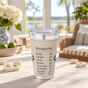 Puede incluir: Un vaso de pl&aacute;stico transparente con tapa y pajita, que presenta una banda de color crema impresa con el texto MAHJONG THE AMERICAN SET y una lista de categor&iacute;as de juego que incluye BAMS, CRAKS, DOTS, WINDS, DRAGONS, FLOWERS, JOKERS y un total de 152.