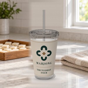 May include: A clear plastic tumbler with a lid and straw, featuring a cream-colored wrap with a dark green four-part floral-style icon, a small gold diamond, and the black text "MAHJONG" followed by "THE TUESDAY FOUR" printed on the front surface.