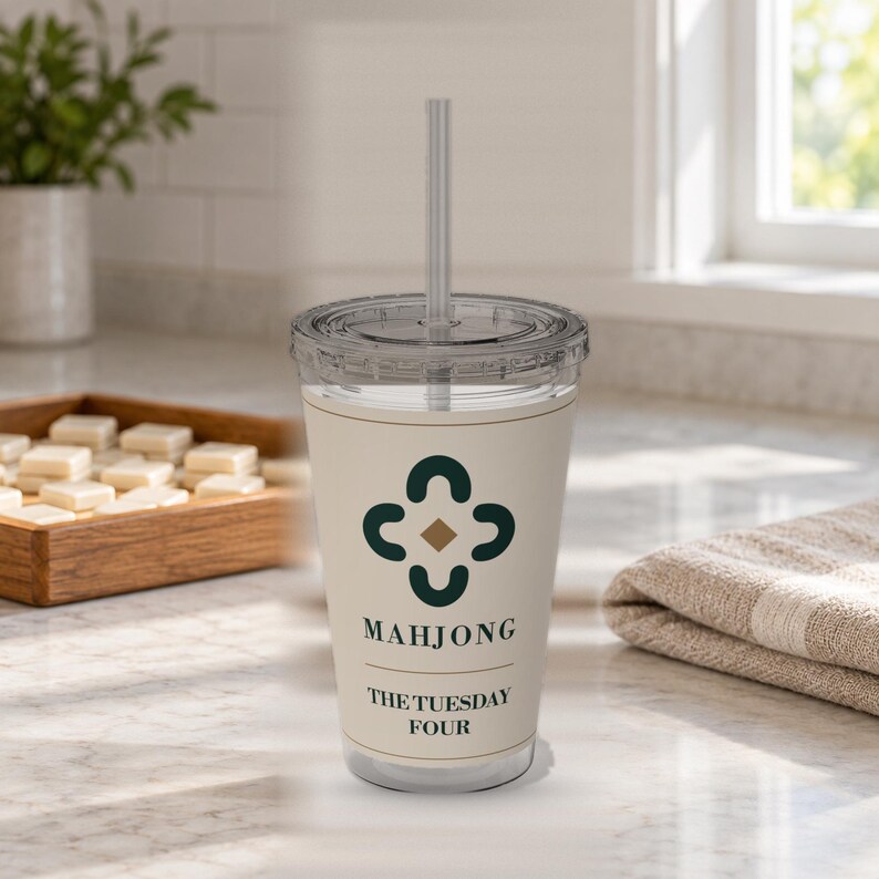 May include: A clear plastic tumbler with a lid and straw, featuring a cream-colored wrap with a dark green four-part floral-style icon, a small gold diamond, and the black text "MAHJONG" followed by "THE TUESDAY FOUR" printed on the front surface.