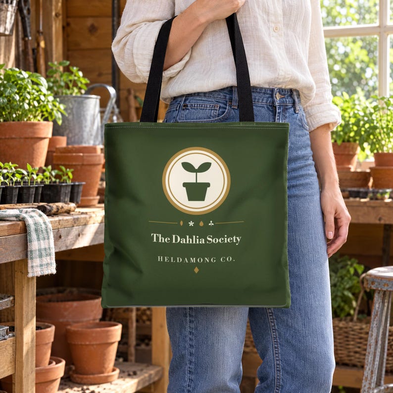 Op de afbeelding: Een donkergroene tote bag met zwarte schouderbanden. De voorzijde is voorzien van een rond logo met een icoon van een plant in een pot, de tekst "The Dahlia Society" en "HELDAMONG CO." in het wit, en kleine decoratieve symbolen.