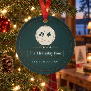 May include: A round dark green ceramic ornament featuring a graphic of two playing cards with club and spade symbols. The text reads The Thursday Four and HELDAMONG CO. in white lettering. A thin gold border circles the edge, and a red ribbon is attached.