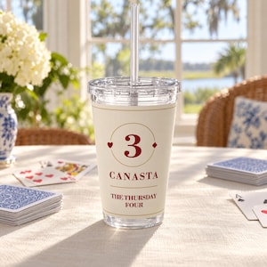 May include: A clear plastic tumbler with a lid and straw, featuring a cream-colored wrap with the red text '3', 'CANASTA', and 'THE THURSDAY FOUR' inside a circular gold border, accented by small red heart and diamond card suit symbols.