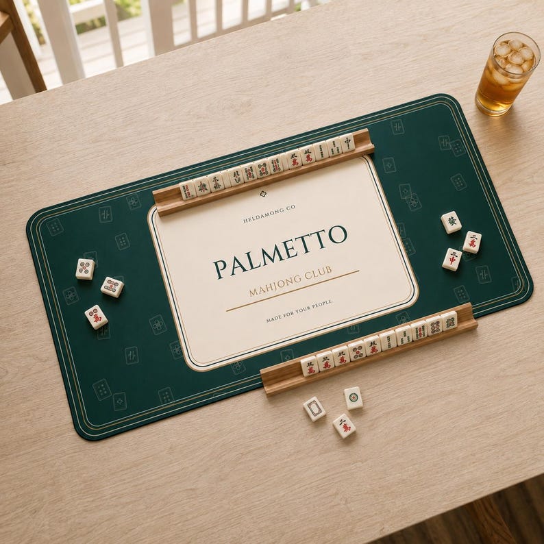May include: A rectangular dark green mahjong mat with a cream-coloured centre featuring the text PALMETTO MAHJONG CLUB. Two wooden tile racks hold white mahjong tiles with red and black characters. Loose tiles are scattered on the mat surface.