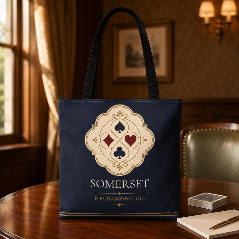 May include: A navy blue tote bag with black handles featuring a central cream-coloured emblem containing black spade and club symbols, a red heart, and a red diamond. Below the emblem, the text reads SOMERSET and HELDAMONG CO. in white lettering.