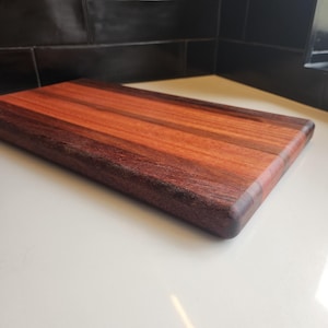 May include: A rectangular wooden chopping board with a two-tone colour scheme. It features alternating stripes of reddish-brown and dark brown wood, with rounded corners. The board is approximately 30 cm long and 20 cm wide.