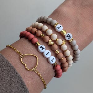 Op de afbeelding: Een verzameling armbanden met kralen in roze, bruine en beige tinten. Sommige armbanden hebben witte letterkralen met de letters "WIT" en "WM". Een gouden hartbedel en een gouden kettingarmband met een hartvormige hanger zijn ook zichtbaar.