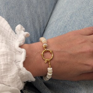 Op de afbeelding: Een armband met kleine, platte, ronde kralen in tinten wit, crème en lichtbruin. De kralen zijn aan elkaar geregen en verbonden door een opvallende, ronde sluiting van goudkleurig metaal die het centrale middelpunt van het sieraad vormt.