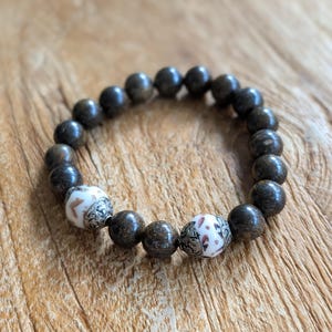 Op de afbeelding: Een armband met donkerbruine, ronde kralen en twee witte sierkralen. De witte kralen hebben een patroon in rood en zwart. De armband wordt getoond op een houten oppervlak.