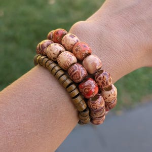 Op de afbeelding: Drie houten armbanden gestapeld op een pols. De bovenste twee armbanden hebben grote, ronde kralen met ingewikkelde patronen en rode accenten. De onderste armband heeft kleinere, olijfgroene, cilindrische kralen. Een natuurlijk, aards accessoire.