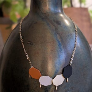 Op de afbeelding: Een gouden ketting met vijf geometrische hangers in bruin, wit en zwart. Elke hanger heeft een gouden druppelvormige bedel. De ketting wordt getoond op een donkere, getextureerde vaas.