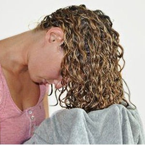May include: A woman with long, curly, light brown hair is shown with her head bent down, her hair covering her face. Her hair is wet and appears to be freshly washed.