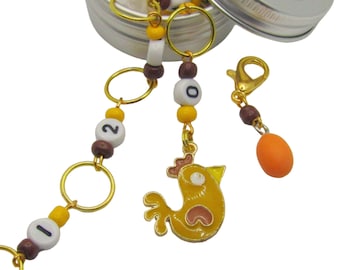 Puede incluir: Una pequeña lata de plata con una tapa transparente que contiene una cadena dorada con cuentas marrones y amarillas, un pequeño amuleto de pollito amarillo y un pequeño amuleto naranja. La cadena tiene cuentas blancas con los números 1, 2 y 0.