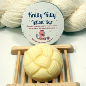 May include: A round, white lotion bar with a braided design sits on a wooden soap dish. The bar is labeled "Knitty Kitty Lotion Bar" and lists the ingredients: beeswax, coconut oil, shea butter, cocoa butter, and lanolin. The label also states that the lotion bar is unscented.