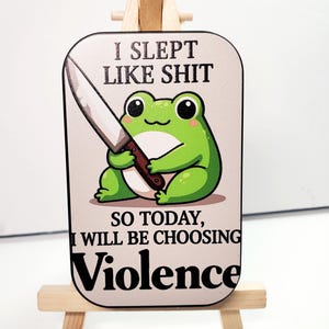 May include: A rectangular sign featuring a cartoon green frog holding a knife. The sign has the text "I SLEPT LIKE SHIT, SO TODAY, I WILL BE CHOOSING VIOLENCE" in black font. The sign is displayed on a small wooden easel.