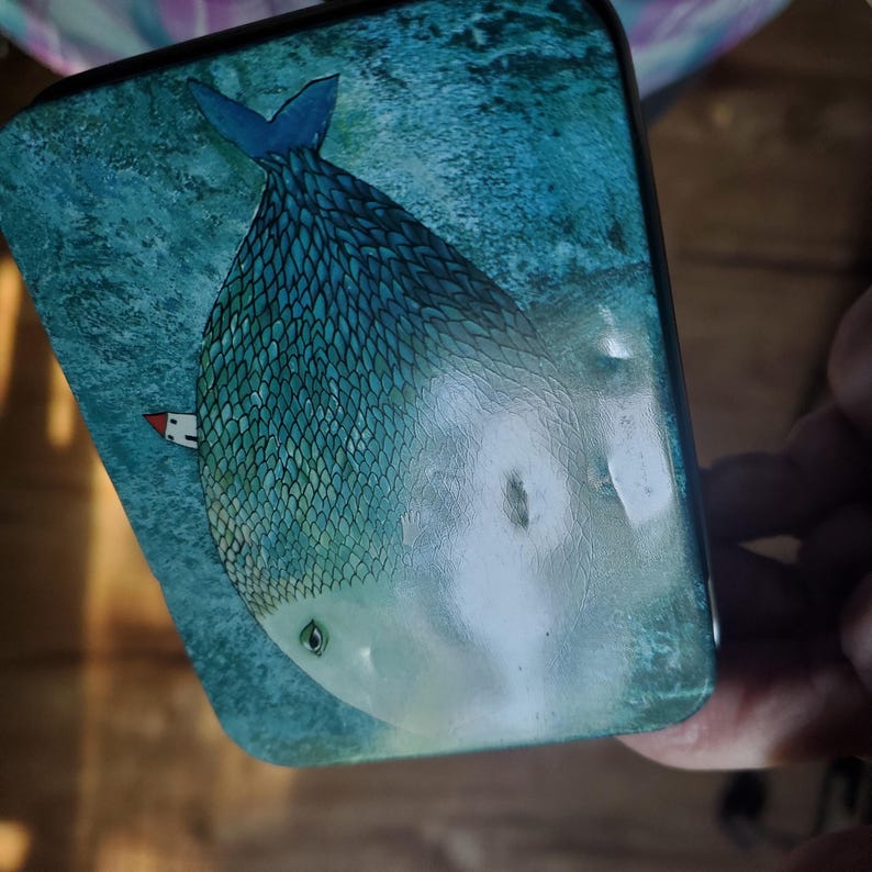 Puede incluir: Una lata met&aacute;lica rectangular con una ilustraci&oacute;n azul y verde de un pez grande con escamas detalladas. El pez tiene un ojo visible y una peque&ntilde;a casa blanca con techo rojo situada a un lado sobre un fondo con textura.