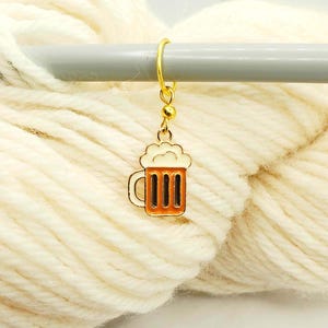 May include: A gold-colored beer mug stitch marker with white foam and a brown beer fill. The charm is hanging from a gold ring, resting on a gray knitting needle, with a background of cream-colored yarn.