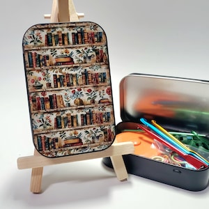 May include: A decorative rectangular tin with a bookshelf design on a small wooden easel. A black tin with crochet hooks and accessories sits beside it. The bookshelf design features rows of books and potted plants.